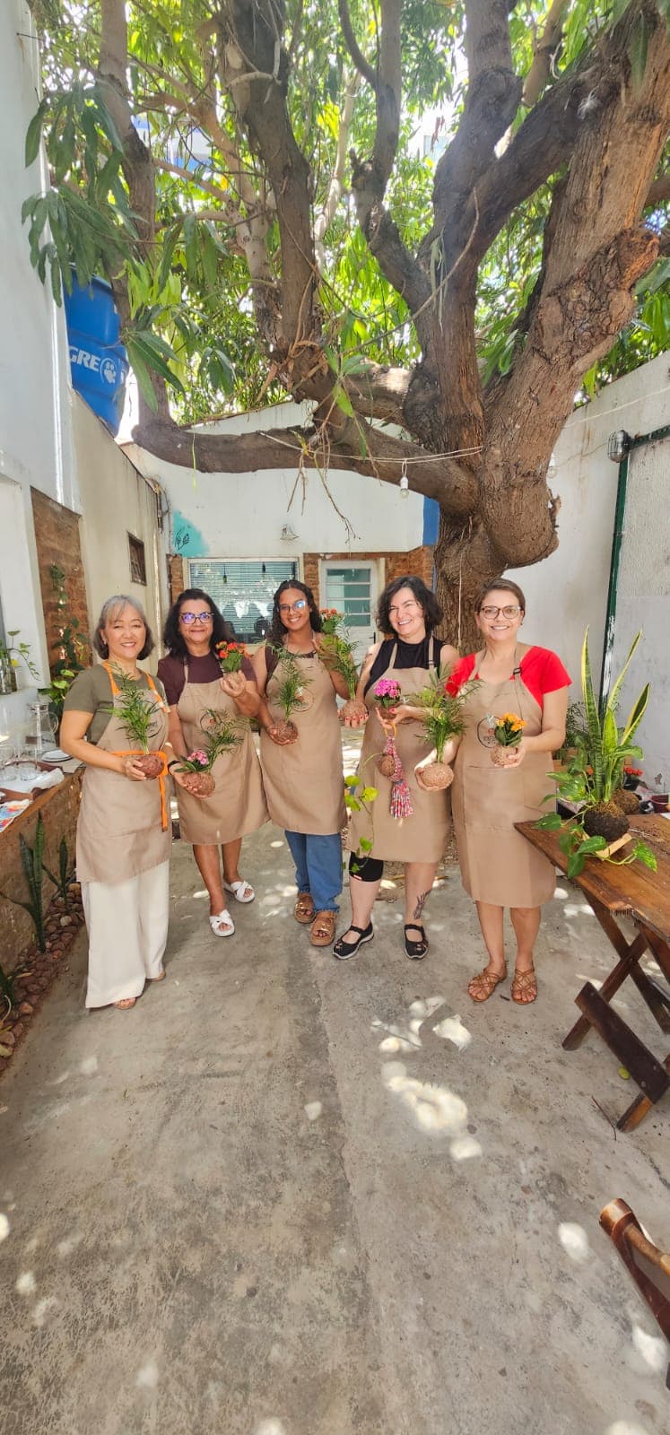 Cinco participantes de avental sob a mangueira, cada uma segurando o kokedama que montou na oficina